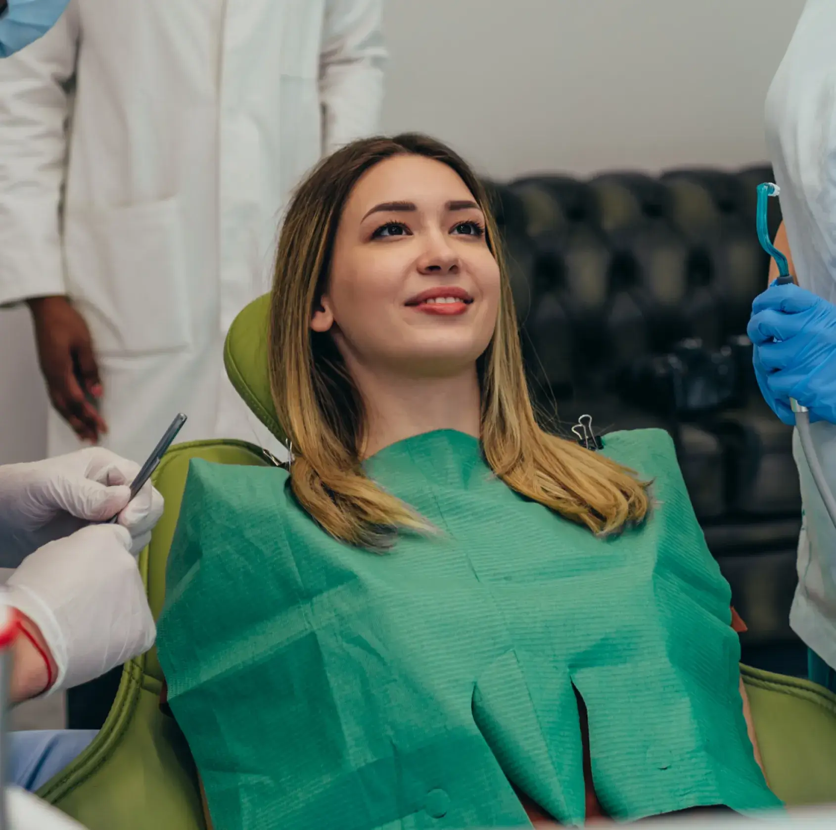 Smiling woman reclining in dental chair with protective bib, surrounded by dental professionals wearing gloves and white coats.