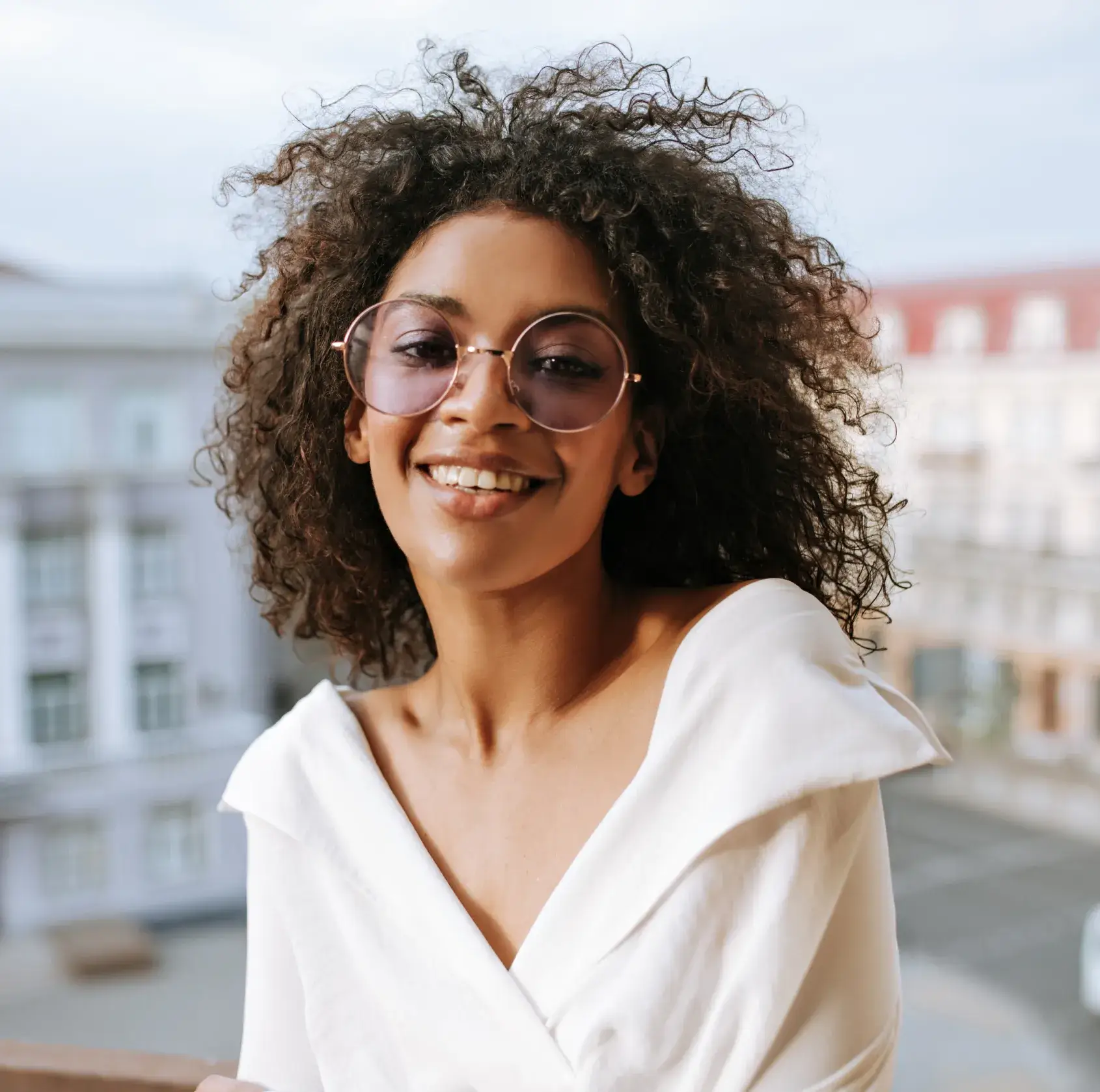 Smiling woman with curly hair wearing round sunglasses and a white off-shoulder top outdoors.