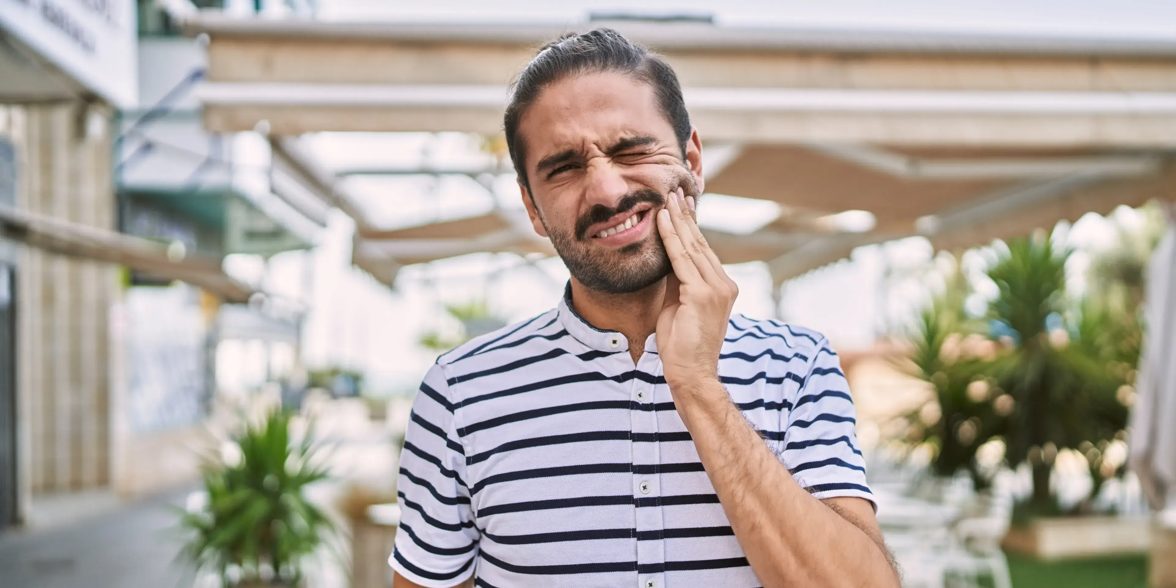 Man in striped shirt holding his cheek with a pained expression, indicating toothache or dental discomfort.