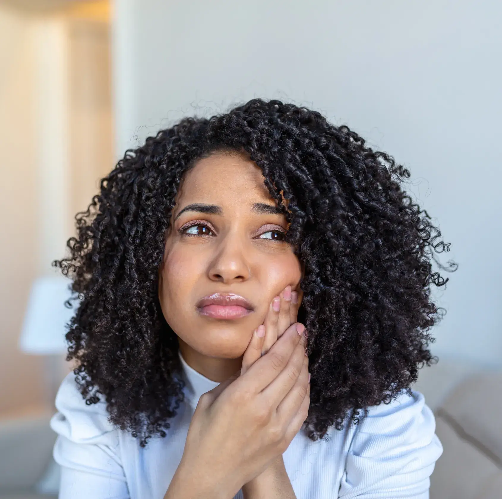 Woman with curly hair holding her cheek with a pained expression, indicating toothache or discomfort.