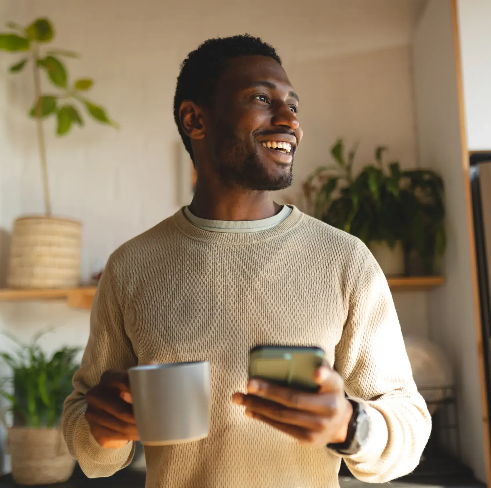Smiling man holding a gray mug and a smartphone indoors with plants in the background.