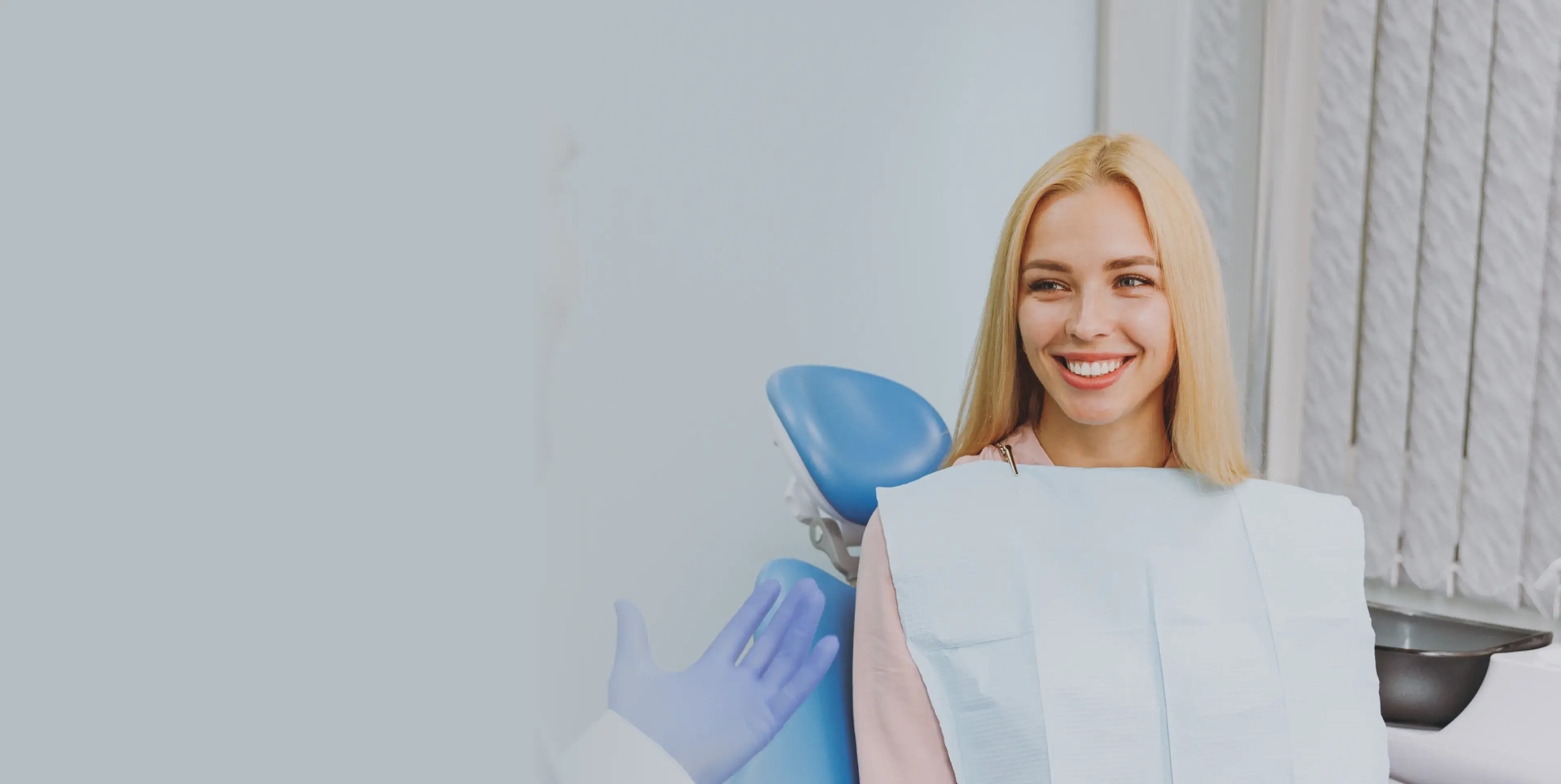 Smiling blonde woman wearing a dental bib seated in a dentist chair while a gloved hand gestures beside her.