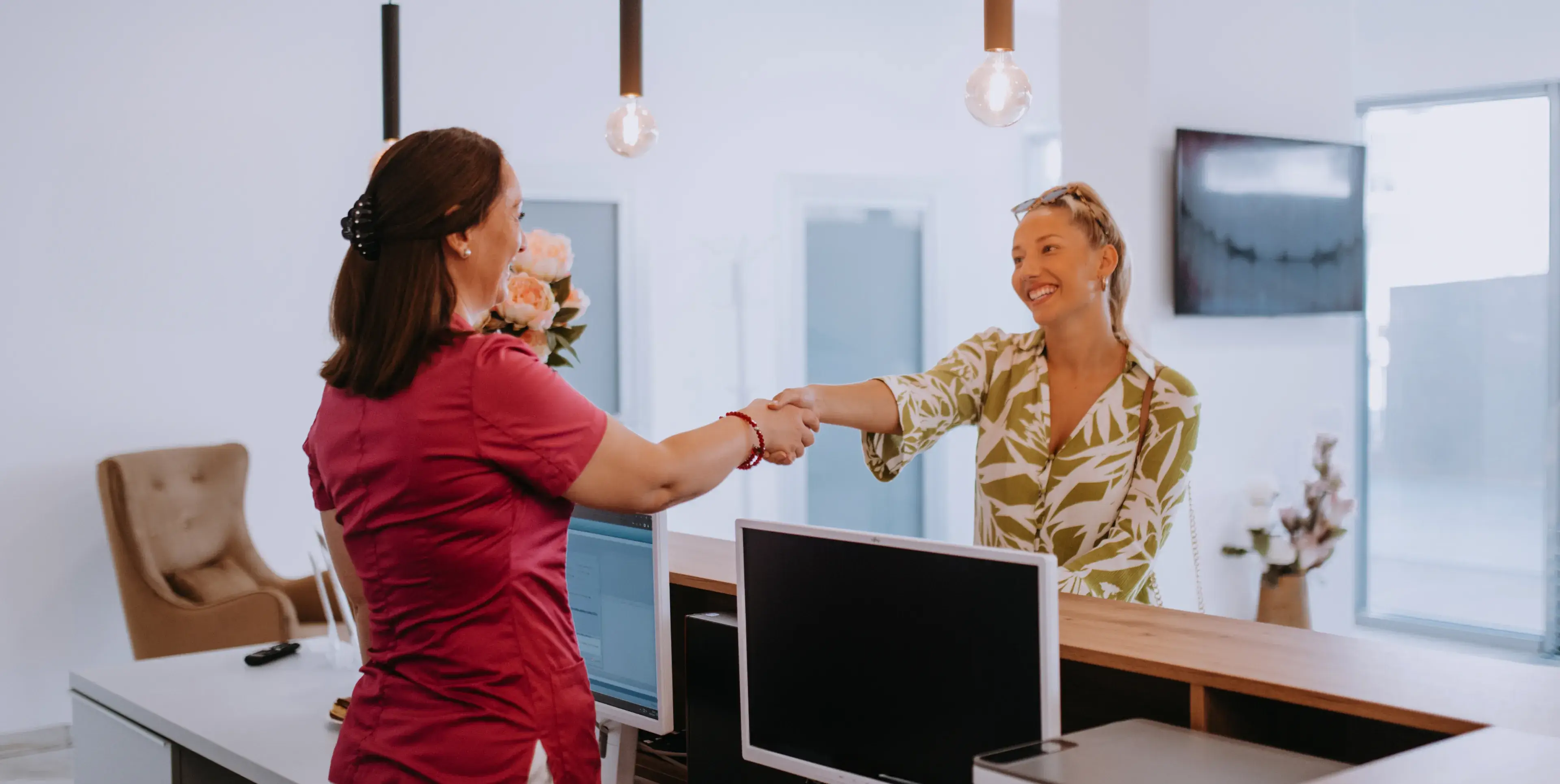 Receptionist in red uniform shaking hands with a smiling blonde woman wearing a green patterned blouse at a modern clinic desk.