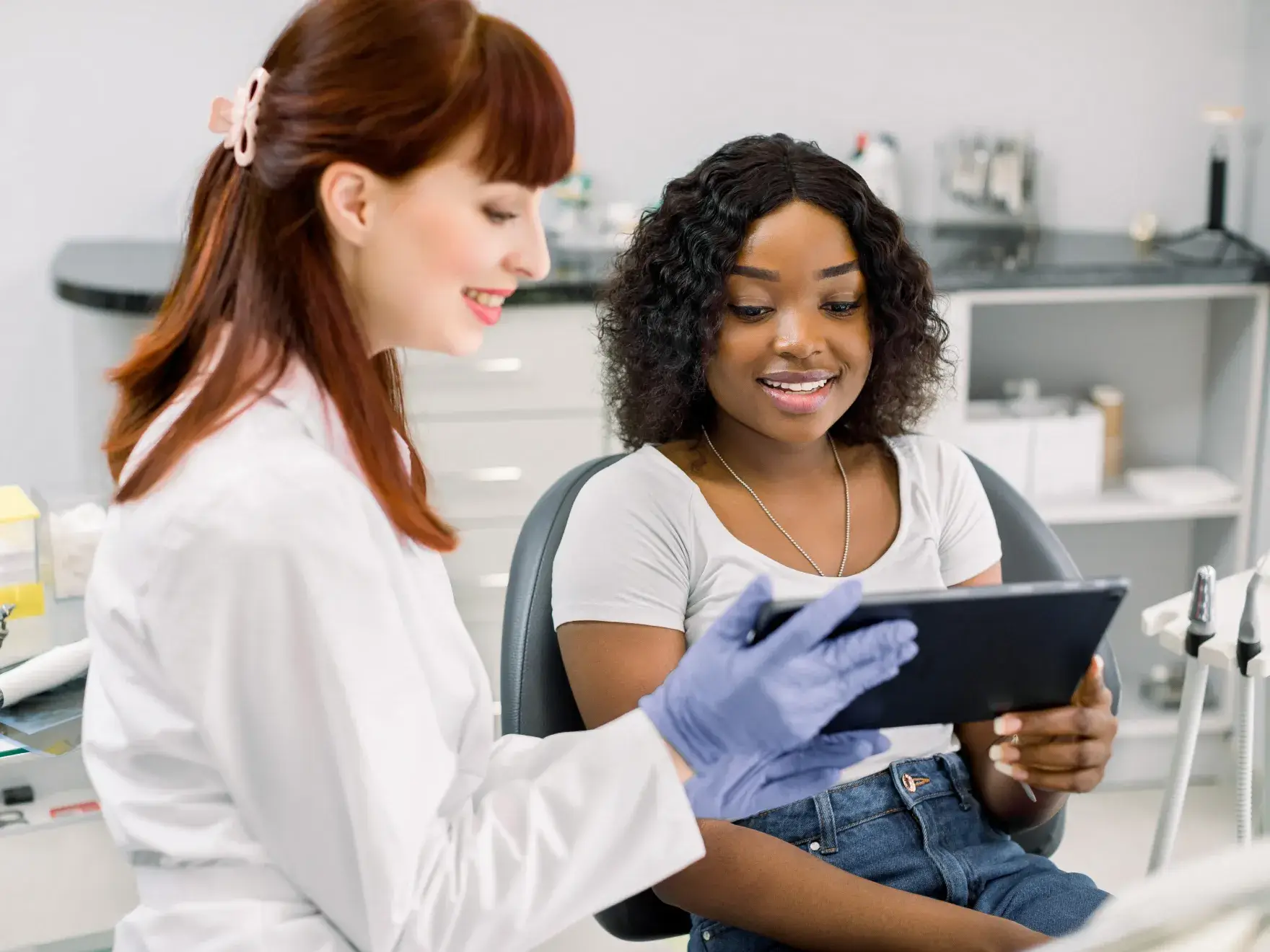 Dentist wearing gloves showing a tablet to a smiling patient sitting in a dental chair.