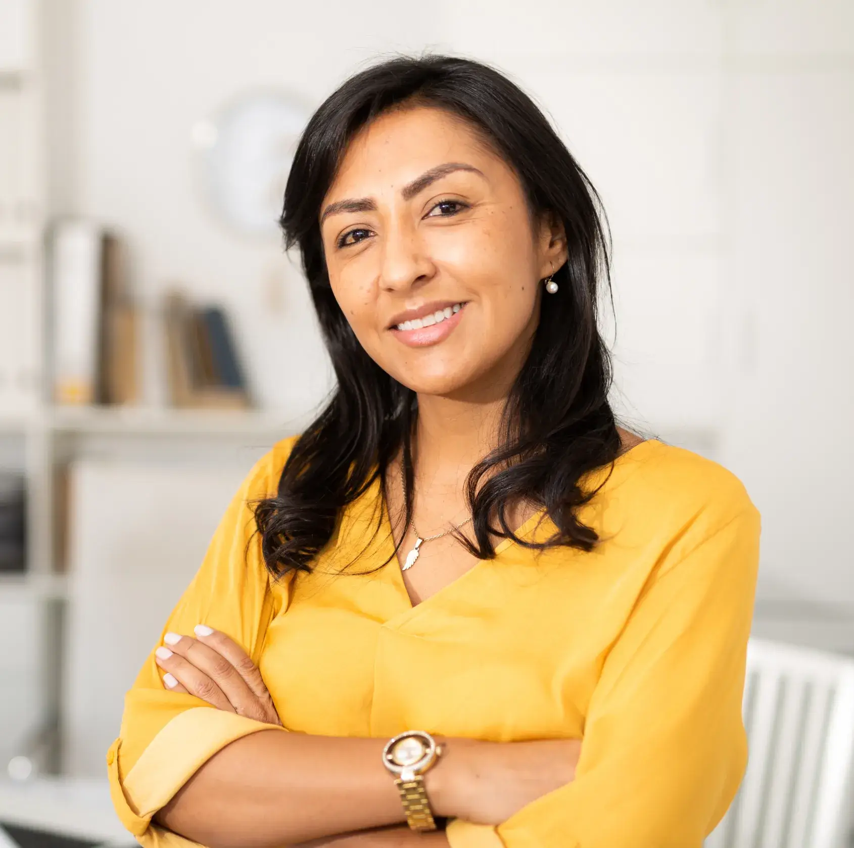Smiling woman with long dark hair, wearing a yellow blouse and gold watch, standing with arms crossed in a bright room.
