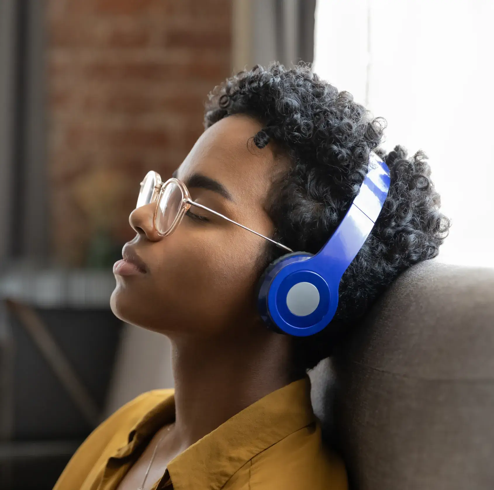 Person with curly hair and glasses leaning back wearing blue headphones and a mustard shirt.