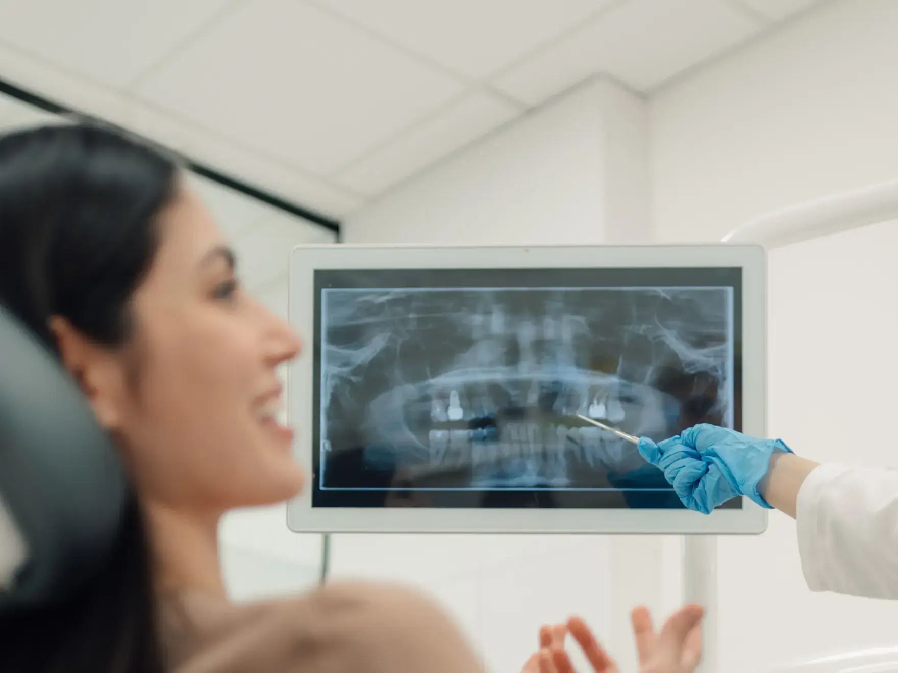 Dentist wearing blue gloves pointing at a dental X-ray on a screen while a female patient smiles in the foreground.