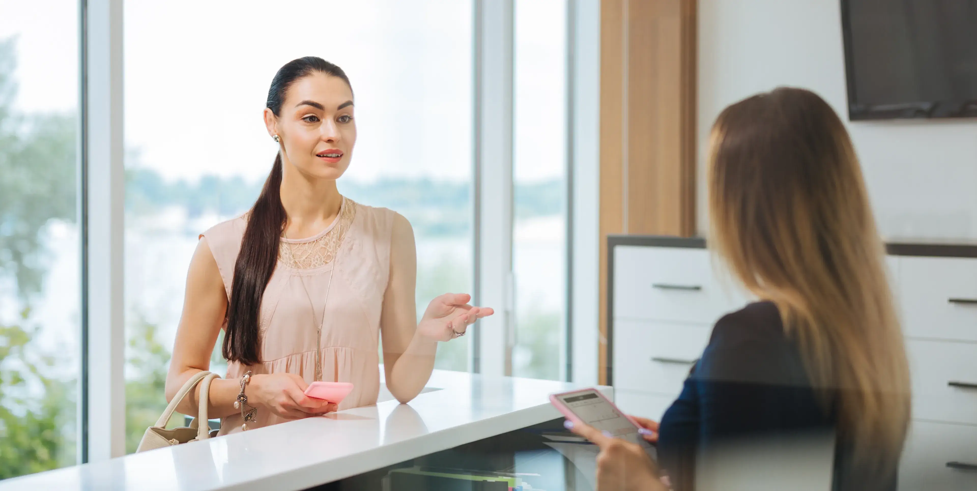 Two women conversing at a bright, modern reception desk holding smartphones, one woman facing the camera in a light pink top.