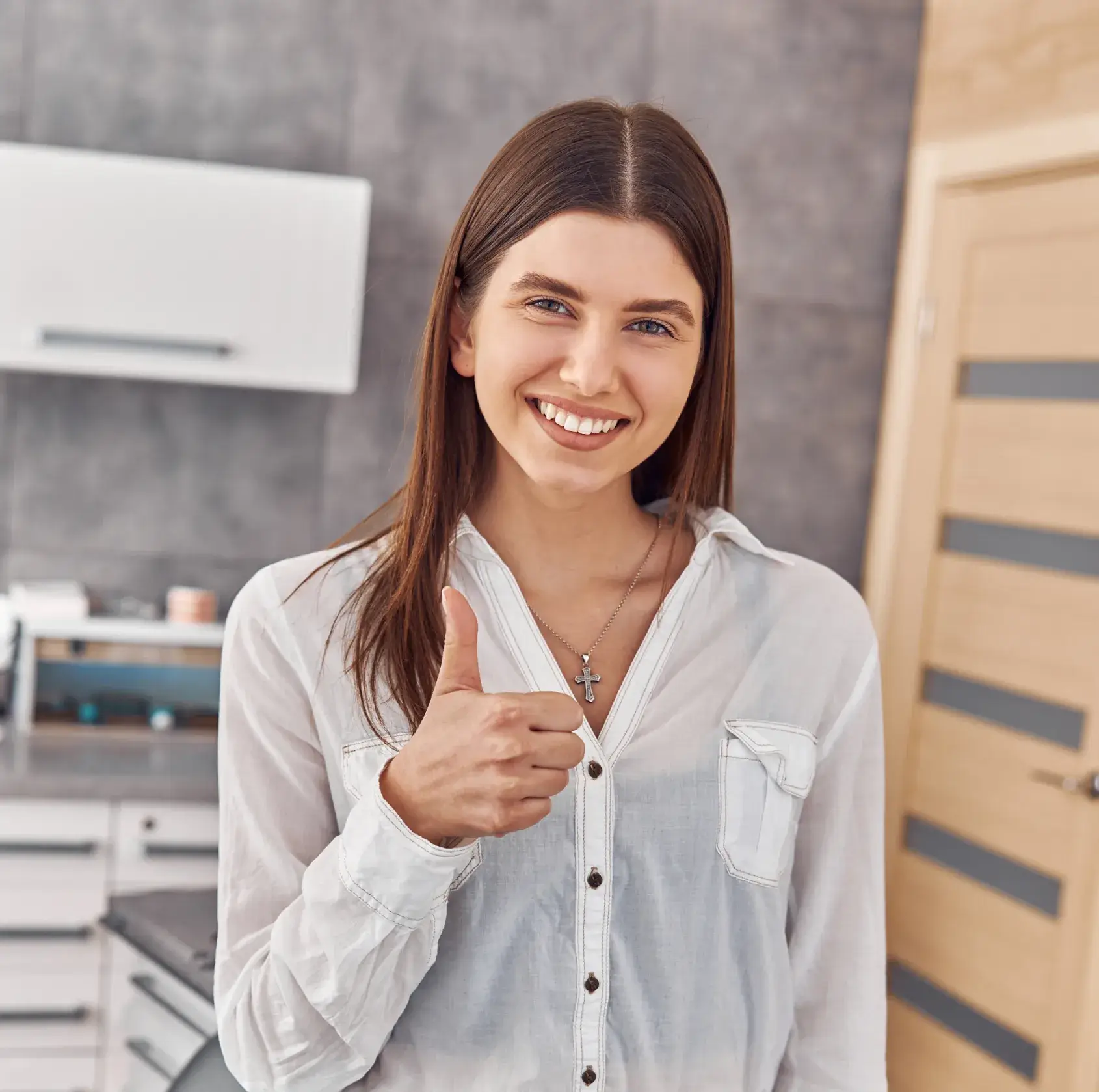 Smiling woman in a white shirt giving a thumbs-up gesture in a modern kitchen.