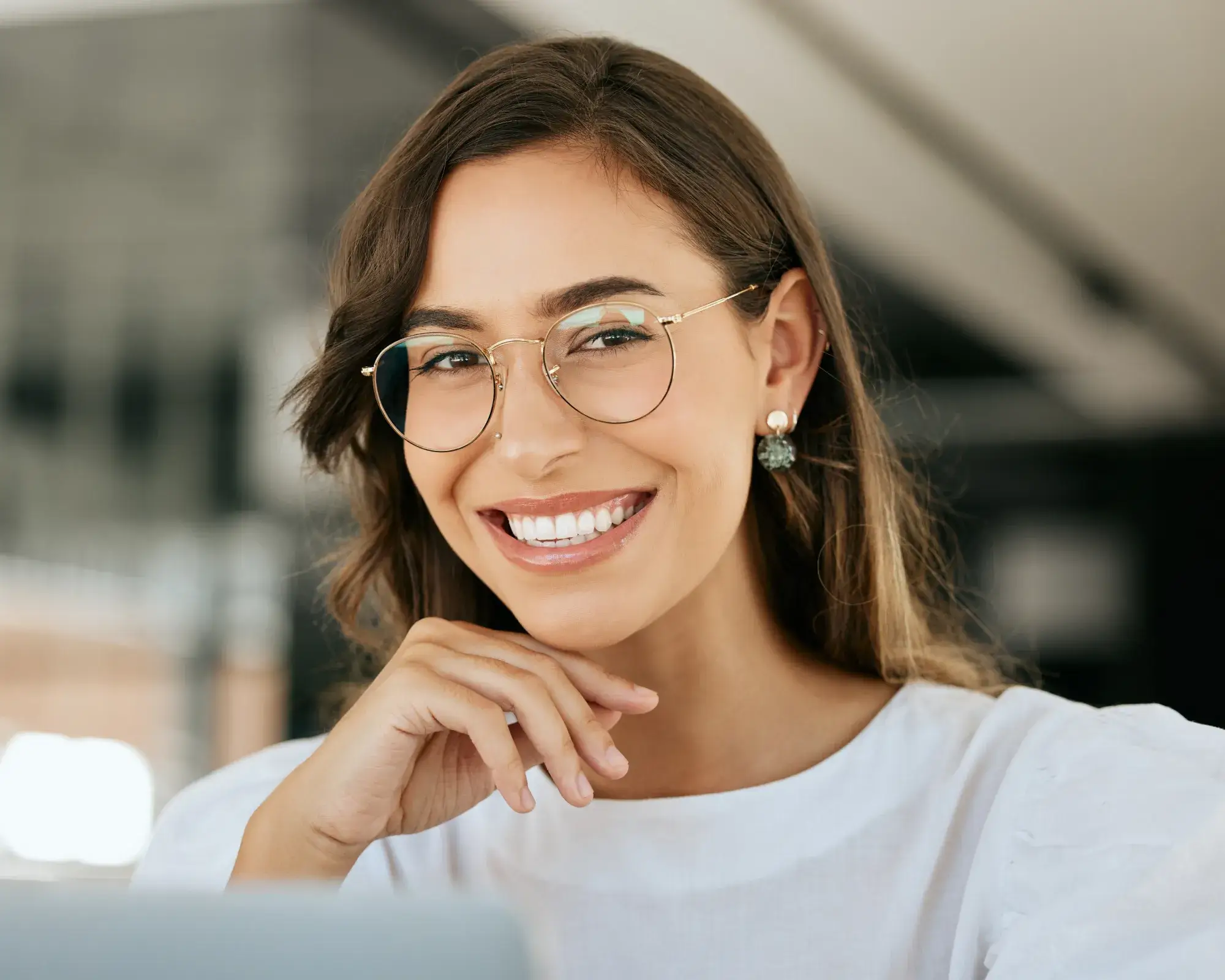 Smiling woman with glasses resting her chin on her hand, wearing white top and earrings.