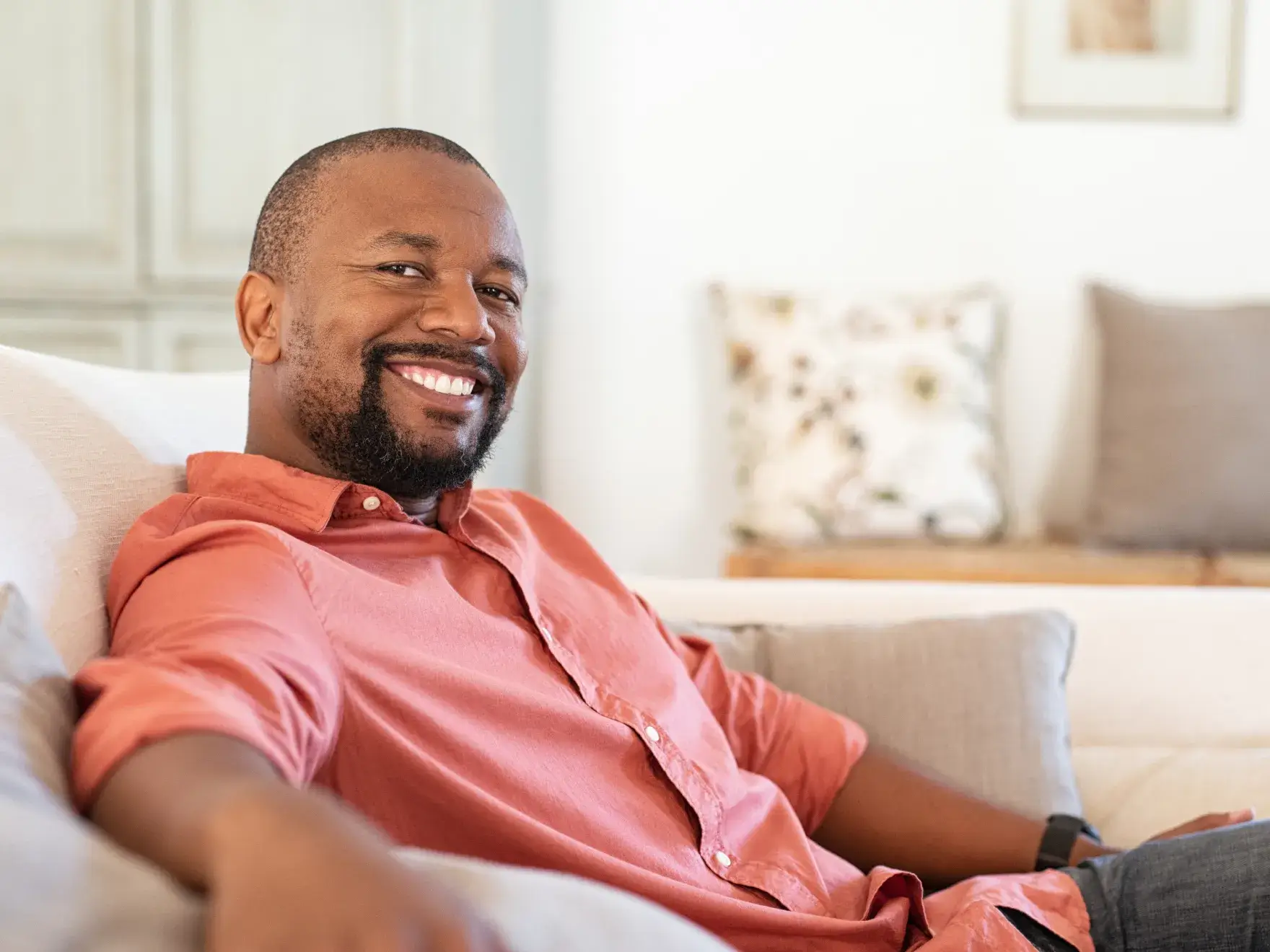 Smiling man in a coral shirt relaxing on a beige sofa in a bright living room.