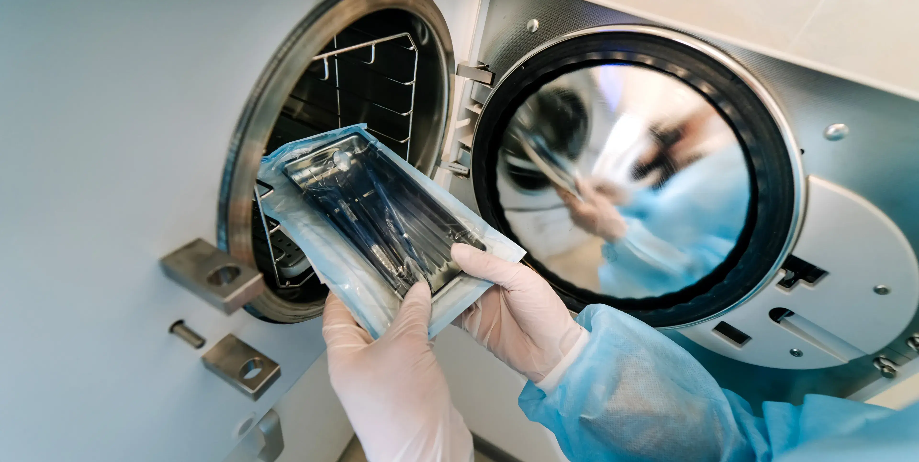 Person wearing gloves and protective clothing holding sealed medical instruments near an open sterilization autoclave.