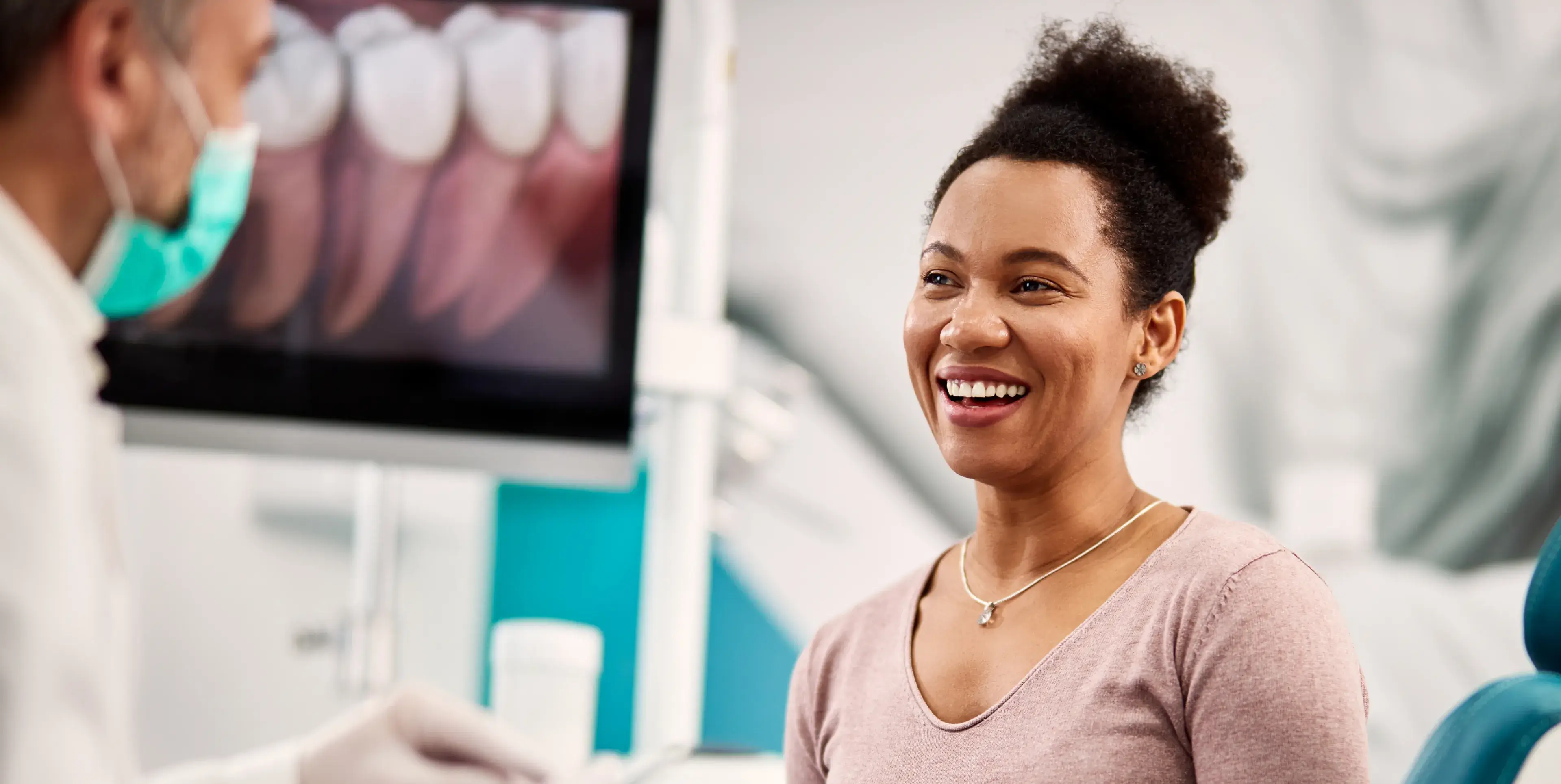 Smiling woman talking with a masked dentist in a dental office with a tooth X-ray on a screen in the background.