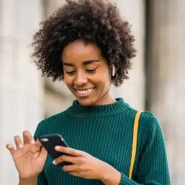 Smiling woman with curly hair in green sweater looking at smartphone outdoors.