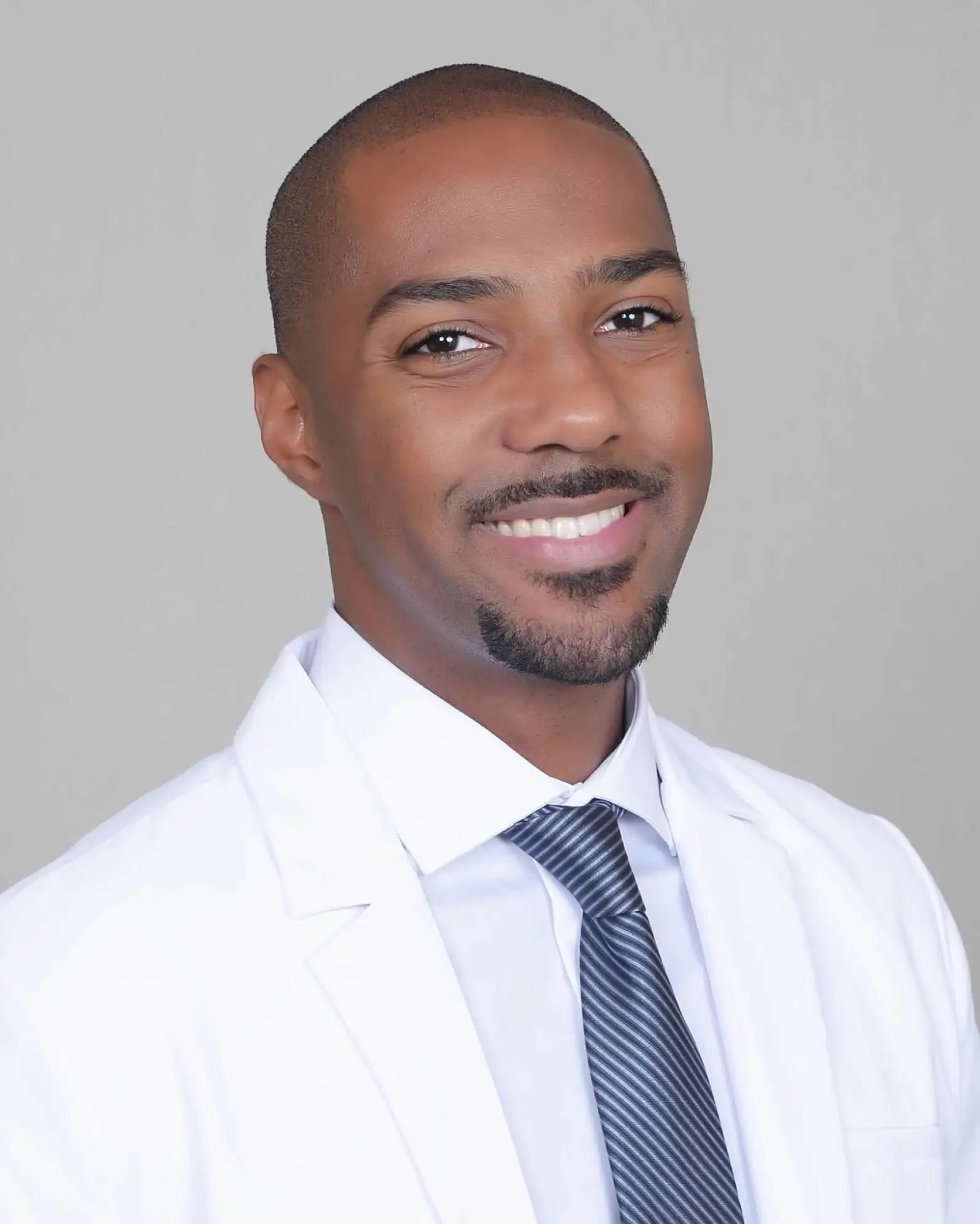 Smiling man wearing a white lab coat, light blue shirt, and striped tie against a plain gray background.