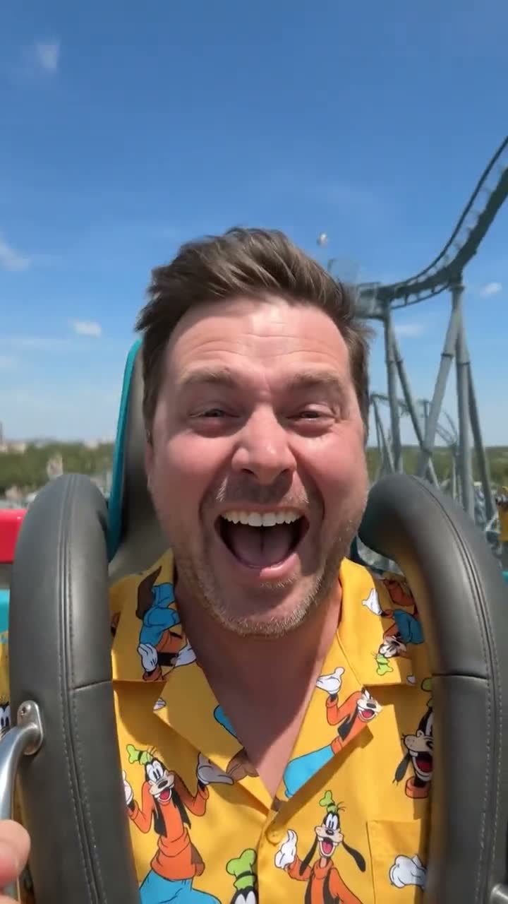 Poster frame from the Playcation vertical film: a man in a yellow Goofy-print Hawaiian shirt laughing on a roller coaster against blue sky