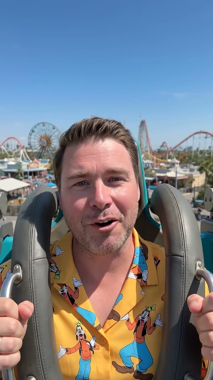 Frame 04, the sign-off: he smiles to camera with the theme park skyline behind him, a Ferris wheel and other coaster tracks in view