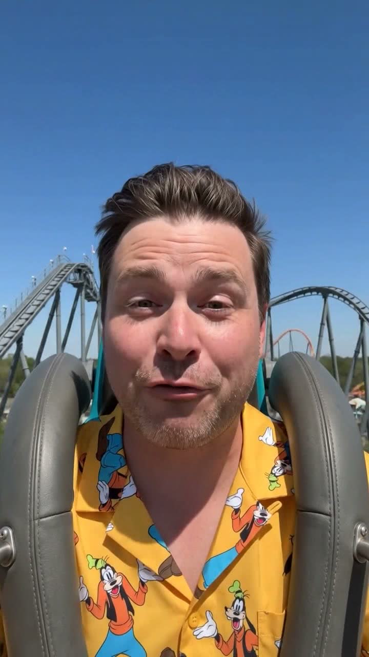 Frame 02, the climb: he talks to camera from the coaster seat as the train climbs the lift hill, track and clear blue sky behind