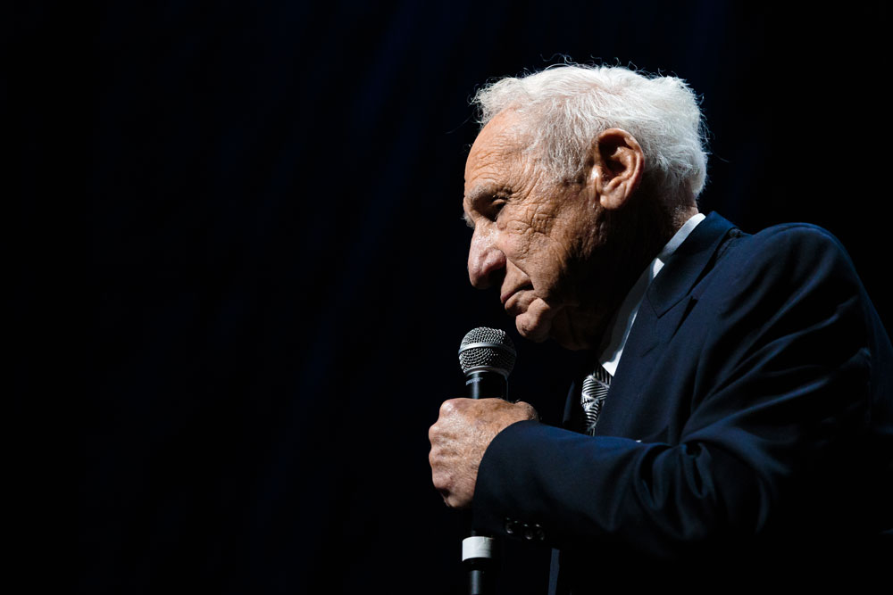 Mel Brooks in profile at the microphone, lit only by the stage against a black background