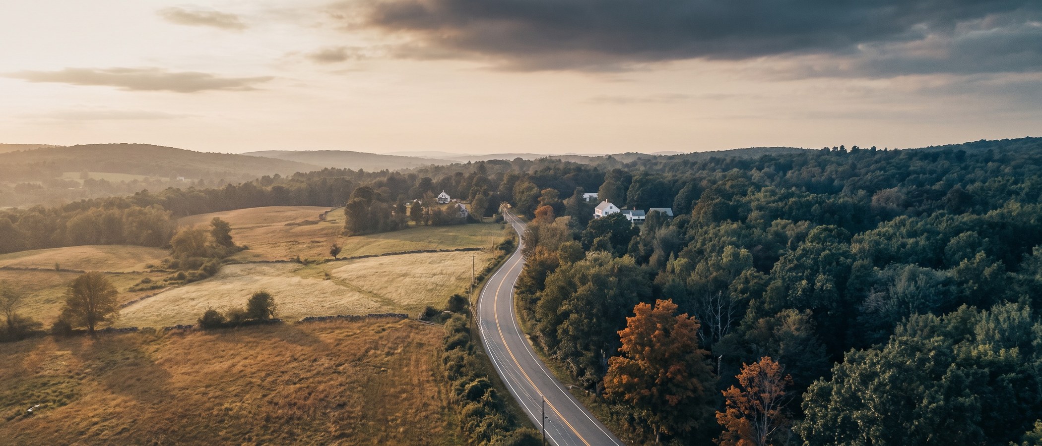 Aerial view of a two-lane road running through the New England countryside at golden hour, with open golden fields on one side and dense green woodland on the other.