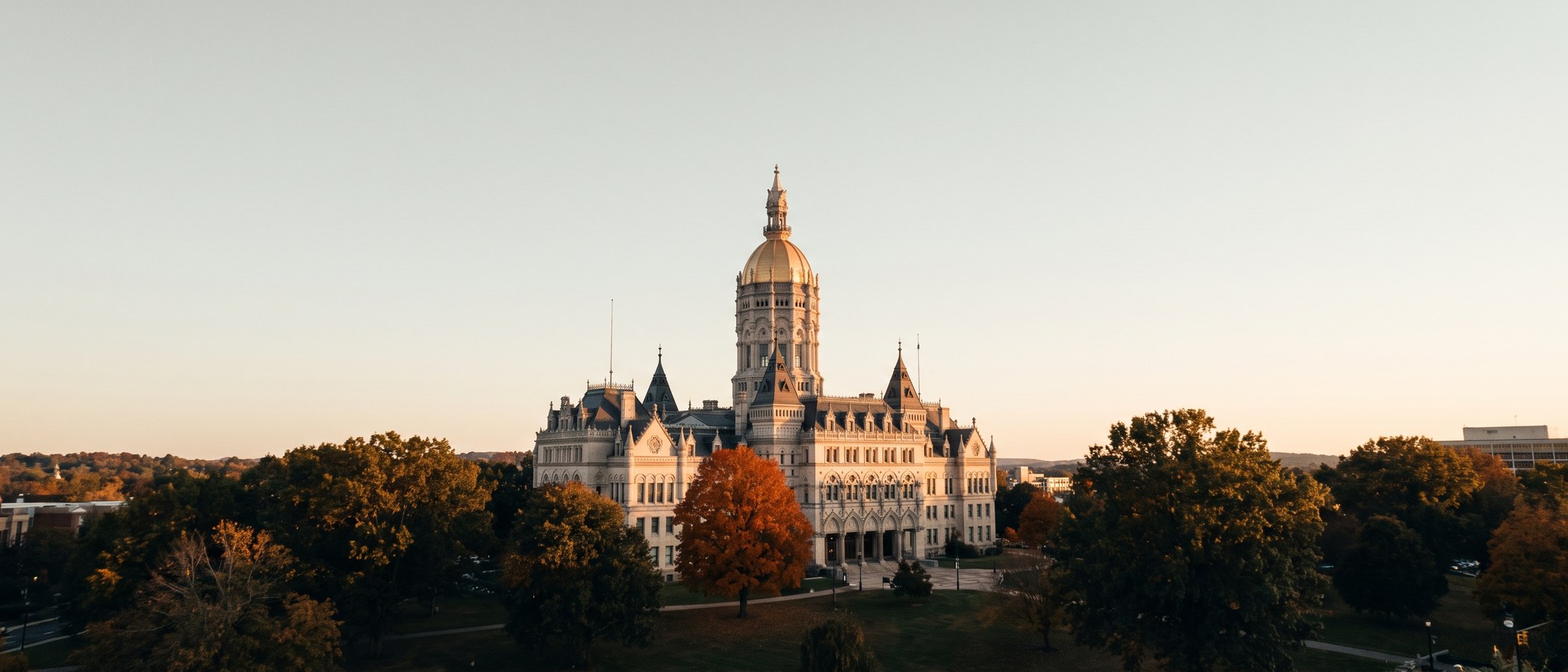 The Connecticut State Capitol building in Hartford at golden hour, framed by trees beginning to turn autumn colors.