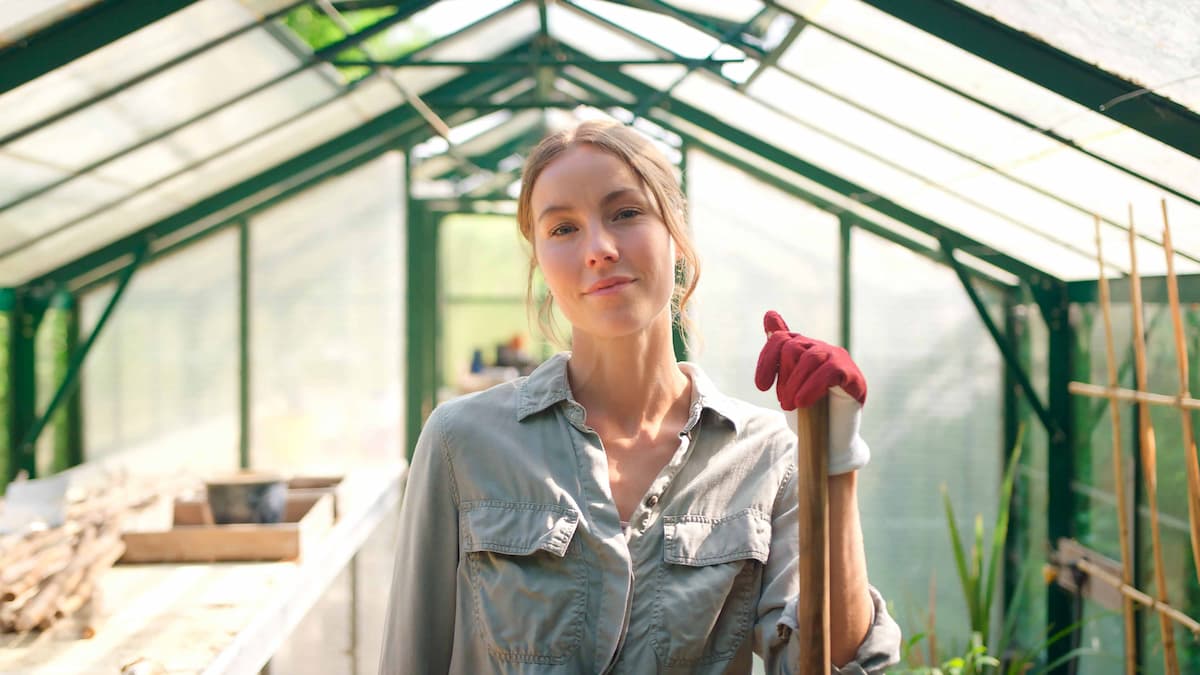 A woman standing inside a greenhouse holding gardening tools
