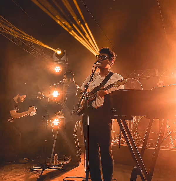 Band performing on stage with two guitarists singing into microphones under warm yellow stage lights.