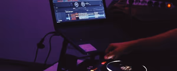 Person adjusting controls on a DJ mixer with a laptop displaying music software in low purple lighting.