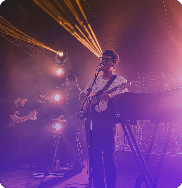 Musicians performing on stage with guitars and keyboards under bright purple and orange stage lights.
