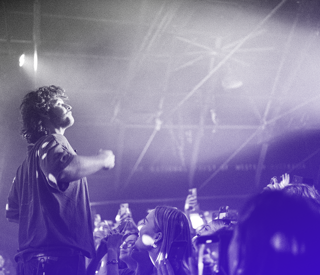 Man singing on stage with audience watching and holding phones in a concert setting.