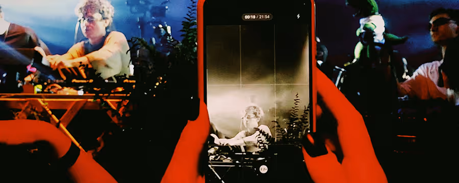 Person in red hands holding a smartphone capturing a photo of a curly-haired individual working on a keyboard in a dimly lit room with plants and other people in the background.