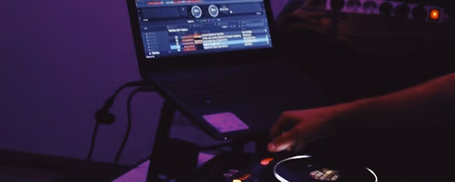 A person using a DJ controller with a laptop displaying music software in a dimly lit setting with purple lighting.