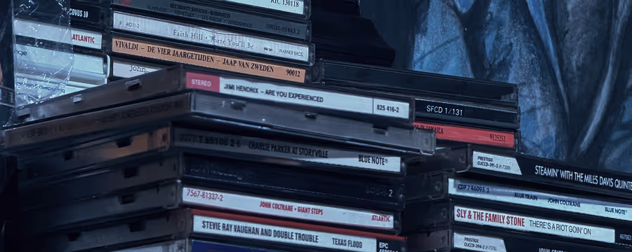 Stacks of vinyl record albums and CD cases featuring various jazz and rock artists, including titles by John Coltrane, Stevie Ray Vaughan, Jimi Hendrix, and Sly & The Family Stone.