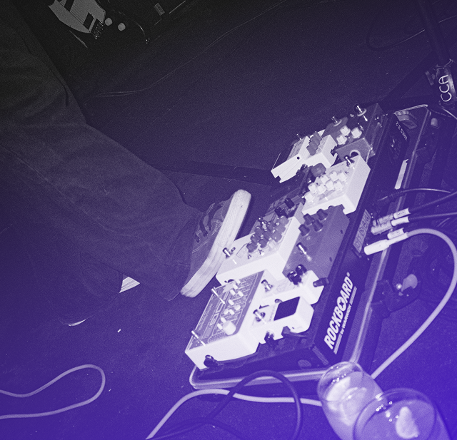 Foot wearing a sneaker operating multiple guitar effect pedals on a Rockboard pedalboard, with cables and two cups nearby.