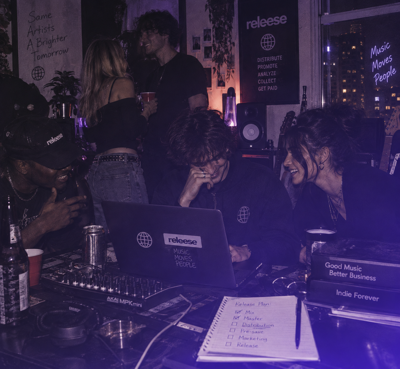 A group of young people in a room with music equipment and laptops branded 'releese.' Two are sitting at a table interacting with a laptop, smiling and talking, while two others stand in the background holding drinks. The table holds a notebook with a checklist titled 'Release Plan' with some tasks checked off, and binders labeled 'Good Music Better Business' and 'Indie Forever.' Posters on the walls include phrases 'Same Artists A Brighter Tomorrow,' 'releese,' and 'Music Moves People.'