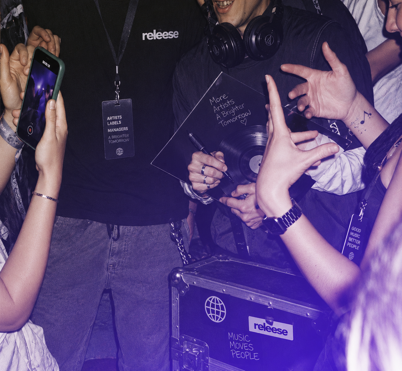 A group of people at a music event, one holding a signed vinyl record with the message 'More Artists A Brighter Tomorrow' and others wearing badges labeled 'ARTISTS LABELS MANAGERS A BRIGHTER TOMORROW' and 'GOOD MUSIC BETTER PEOPLE'. Hands are visible with one person filming on a smartphone and others gesturing. A black equipment case with stickers reading 'releese' and 'Music Moves People' is in the foreground.