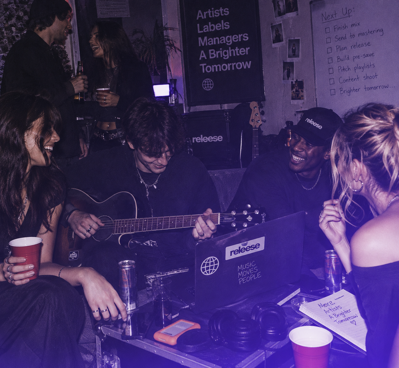 Group of five young adults socializing in a cozy room with music gear; one person is playing acoustic guitar, others are chatting and smiling, some holding drinks. A laptop with a 'releese' sticker and 'Music Moves People' text is on a table cluttered with headphones, cans, and a notebook. Posters on the wall read 'Artists Labels Managers A Brighter Tomorrow' and a whiteboard lists tasks including finishing mix, mastering, planning release, building pre-save, pitching playlists, content shoot, and 'Brighter tomorrow.'
