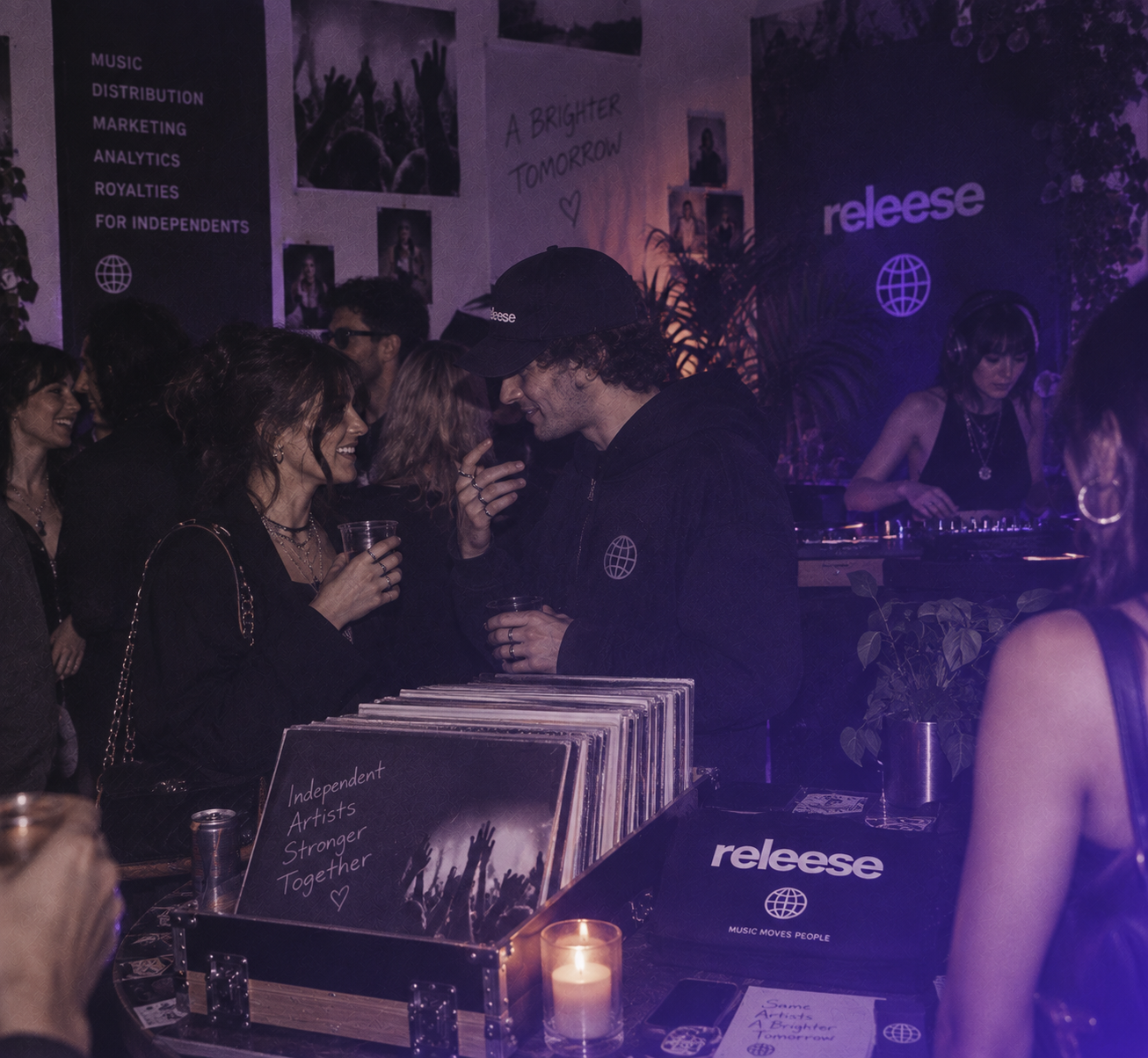 People socializing in a dimly lit venue with purple lighting; a man and woman are holding drinks and talking near a box of vinyl records that has a cover reading "Independent Artists Stronger Together"; in the background, a DJ wearing headphones is playing music at a booth with the word "releese" and a globe logo, and posters on the wall include phrases like "A Brighter Tomorrow" and services such as music distribution and marketing.