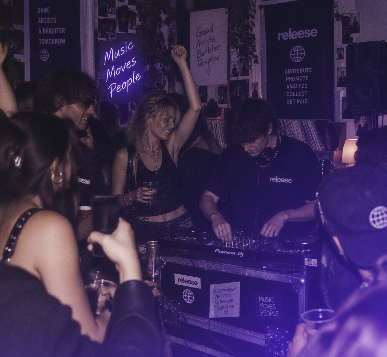 A DJ wearing a 'releese' shirt operates a Pioneer DJ mixer at a crowded indoor party where people dance and hold drinks; neon signs read 'Music Moves People' and 'Independent Artists Stronger Together'.