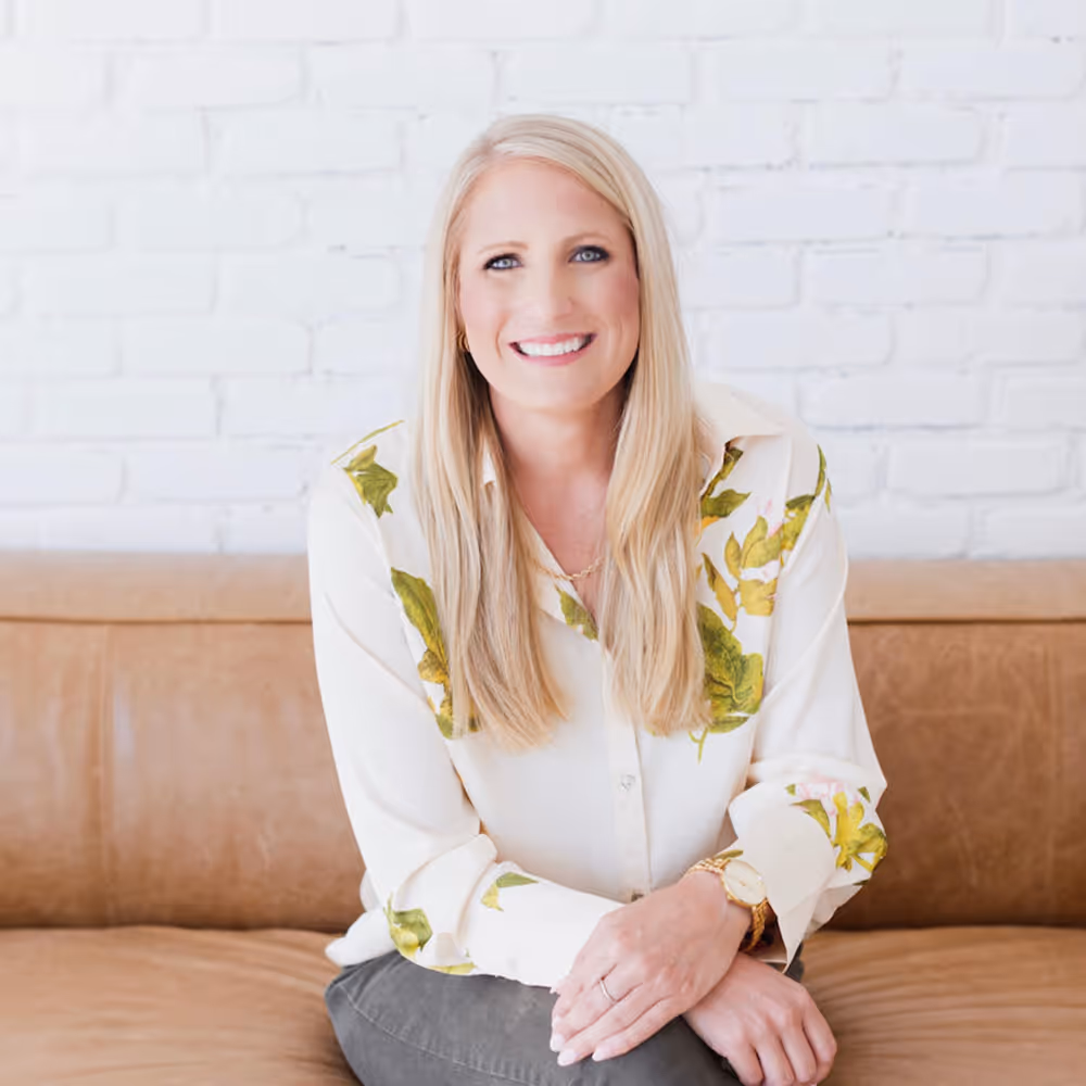 Smiling blonde woman with long hair wearing a white blouse with green leaf patterns, sitting on a tan leather couch against a white brick wall.