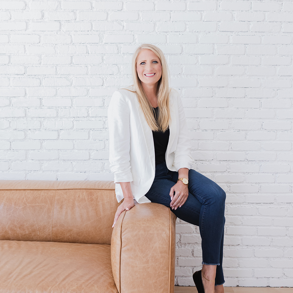 Smiling woman with long blonde hair wearing a white blazer, black top, and jeans sitting on the arm of a tan leather sofa against a white brick wall. Tina Smith, LPC owner of Ray of Hope Counseling. A virtual mental health practice serving texas.