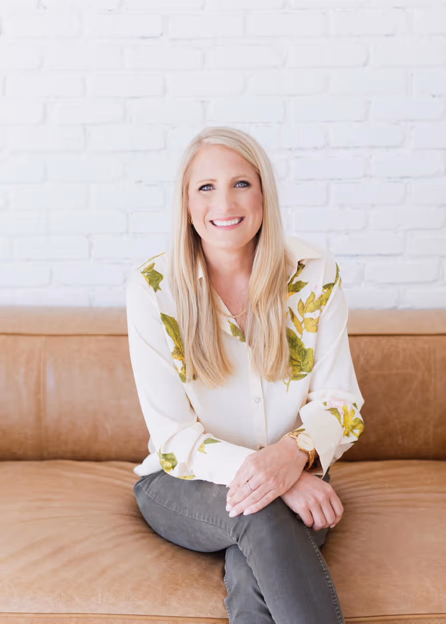 Smiling blonde woman wearing a white blouse with green leaves and gray pants sitting cross-legged on a brown leather couch against a white brick wall.