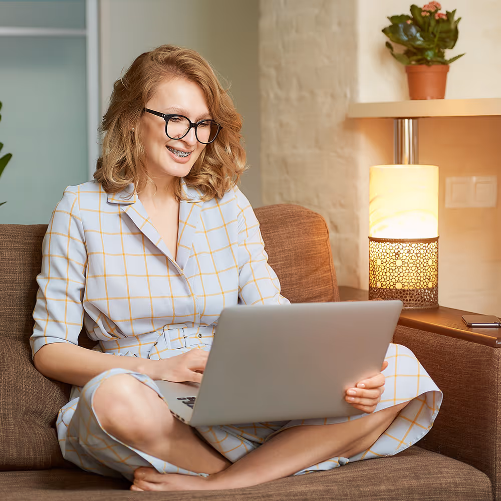 Smiling woman with glasses and braces sitting cross-legged on a brown couch using a laptop.