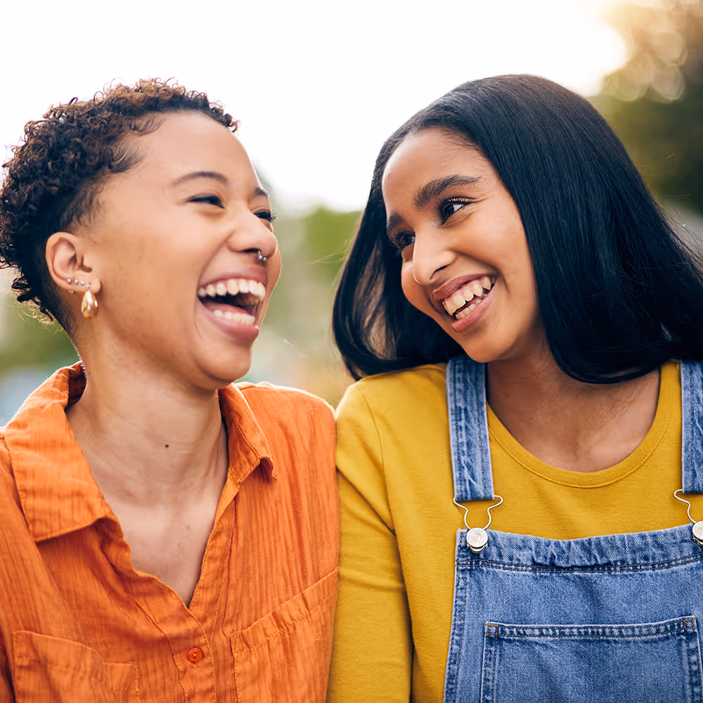 Two young women smiling and laughing warmly at each other outdoors in casual clothing.