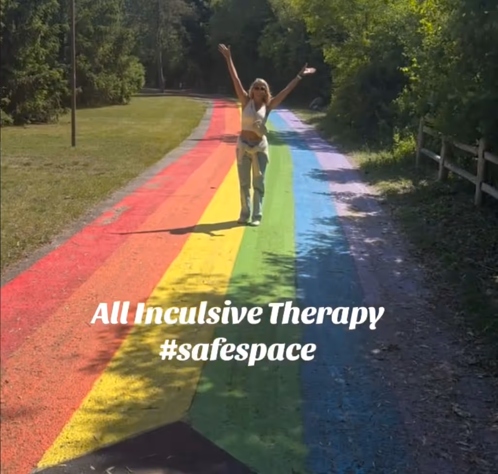 Person with raised arms standing on a rainbow-colored path surrounded by trees and grass, with text ‘All Inclusive Therapy #safespace’ overlaid.