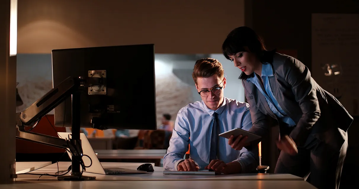 Woman in business suit showing tablet to a man in glasses and tie working on a keyboard in a dimly lit office.
