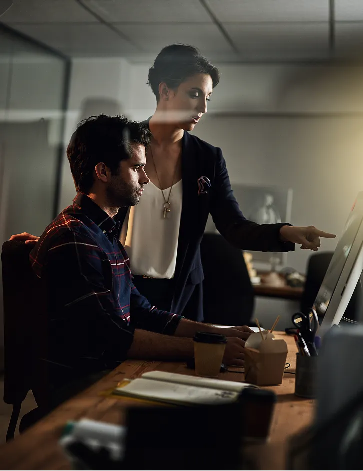 Woman standing and pointing at a computer screen while man sits at desk looking at the screen in a dimly lit office.