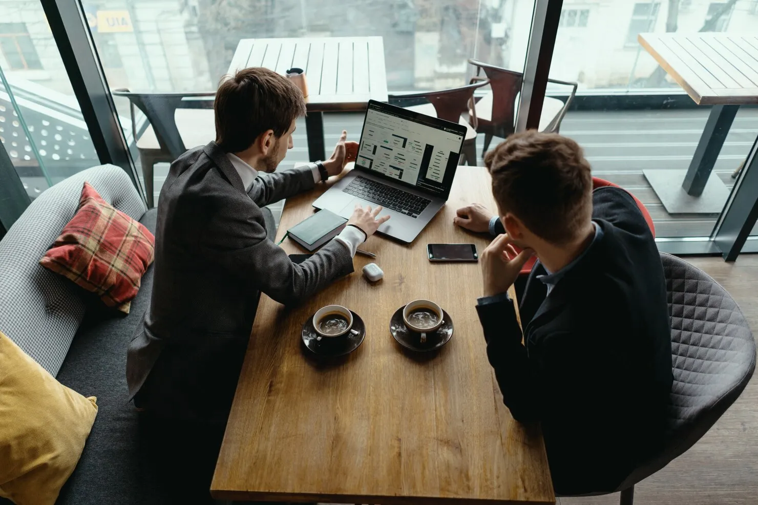 Two men in business attire sitting at a wooden table with coffee cups, looking at financial data on a laptop in a modern cafe with large windows.