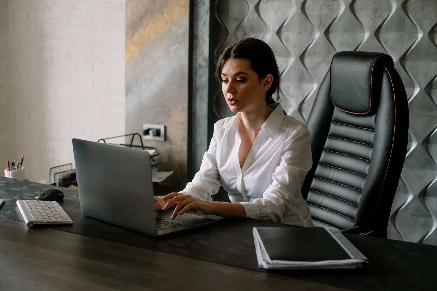 Woman in a white shirt typing on a laptop at a desk with office supplies and folders.