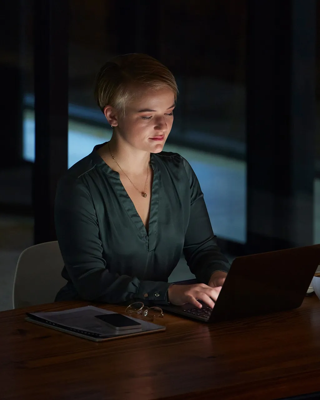 Woman with short hair working on a laptop at a wooden table in a dimly lit room.
